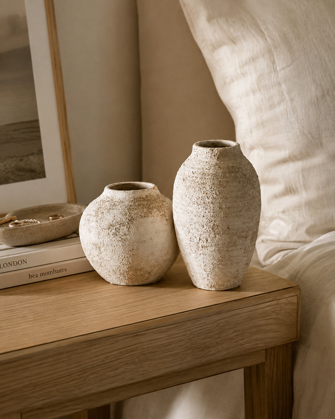 Sand white pumice stone vase pair on oak nightstand with linen cushions, jewellery tray and art books in bedroom