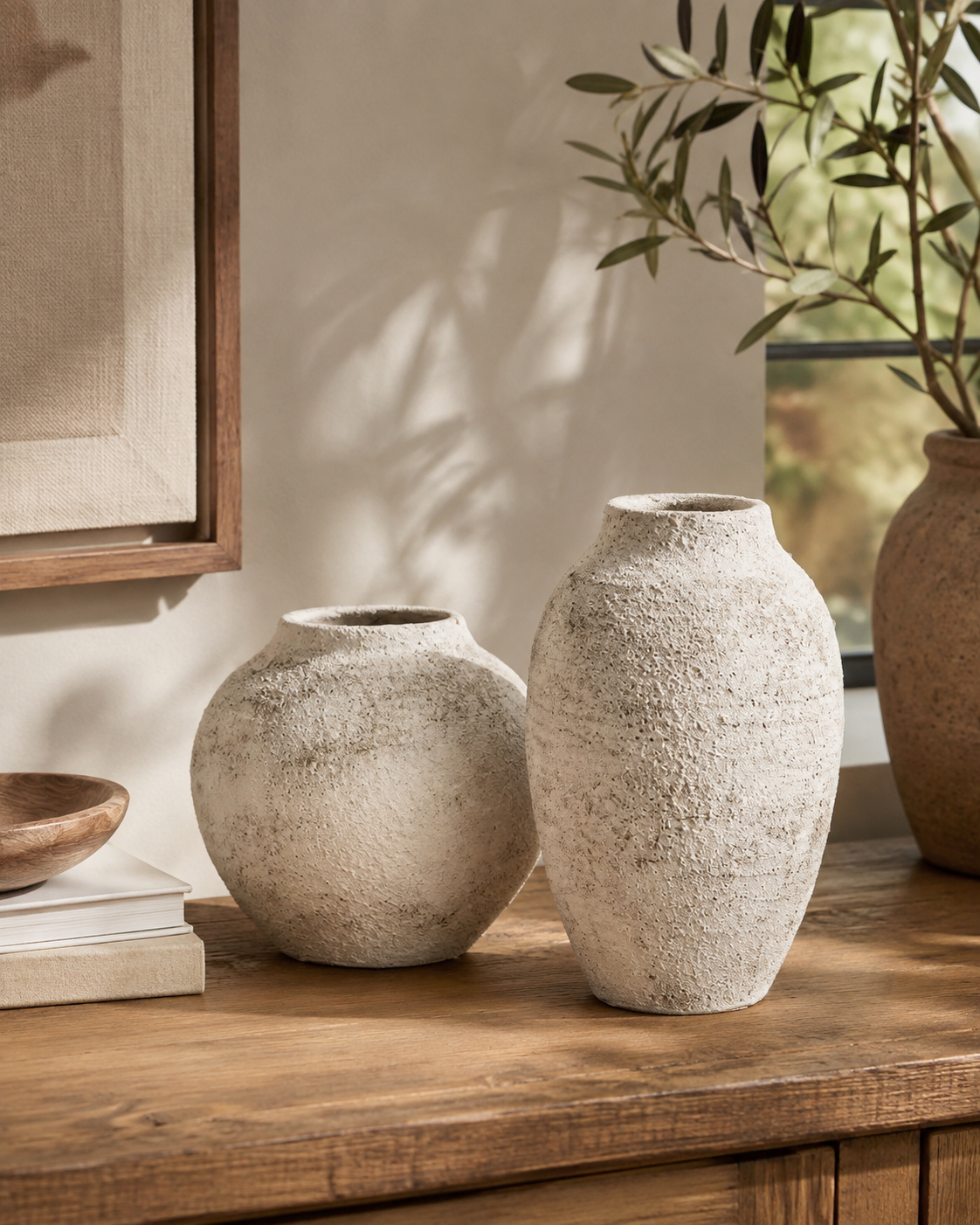 Two raw textured stone vases on oak sideboard beside wood bowl and olive tree in dappled sunlit living room