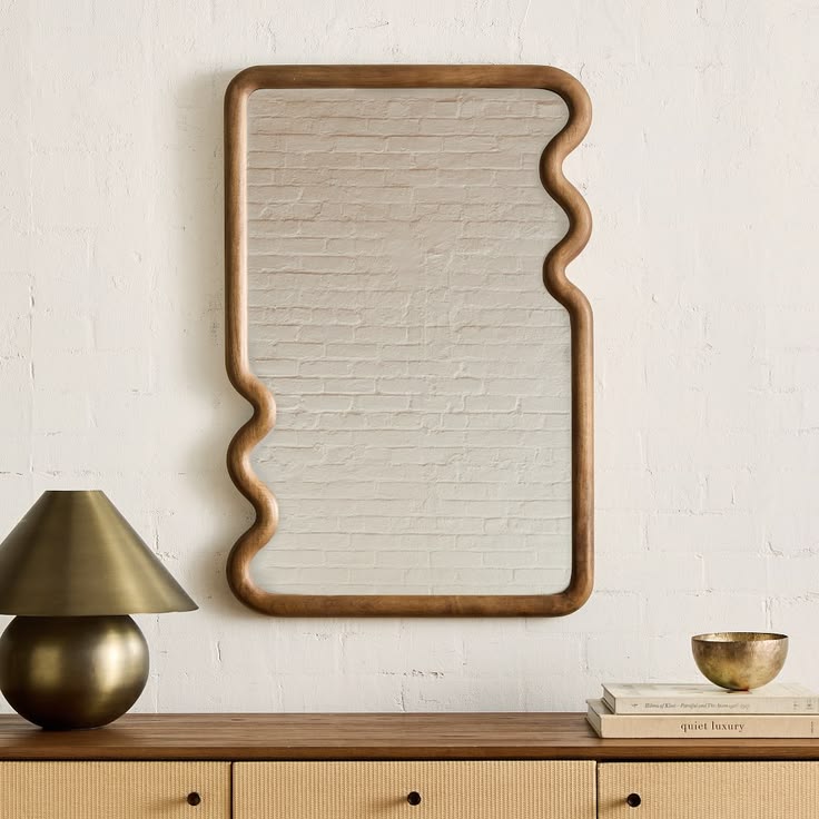 Large wavy wood frame mirror leaning against exposed brick wall above rattan sideboard with brass lamp and gold bowl